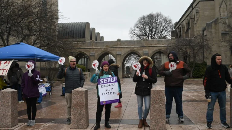 A group of striking people stand in a line on BU's Marsh Plaza in the rain. They are mid-chant and carrying SEIU and BUGWU signs.